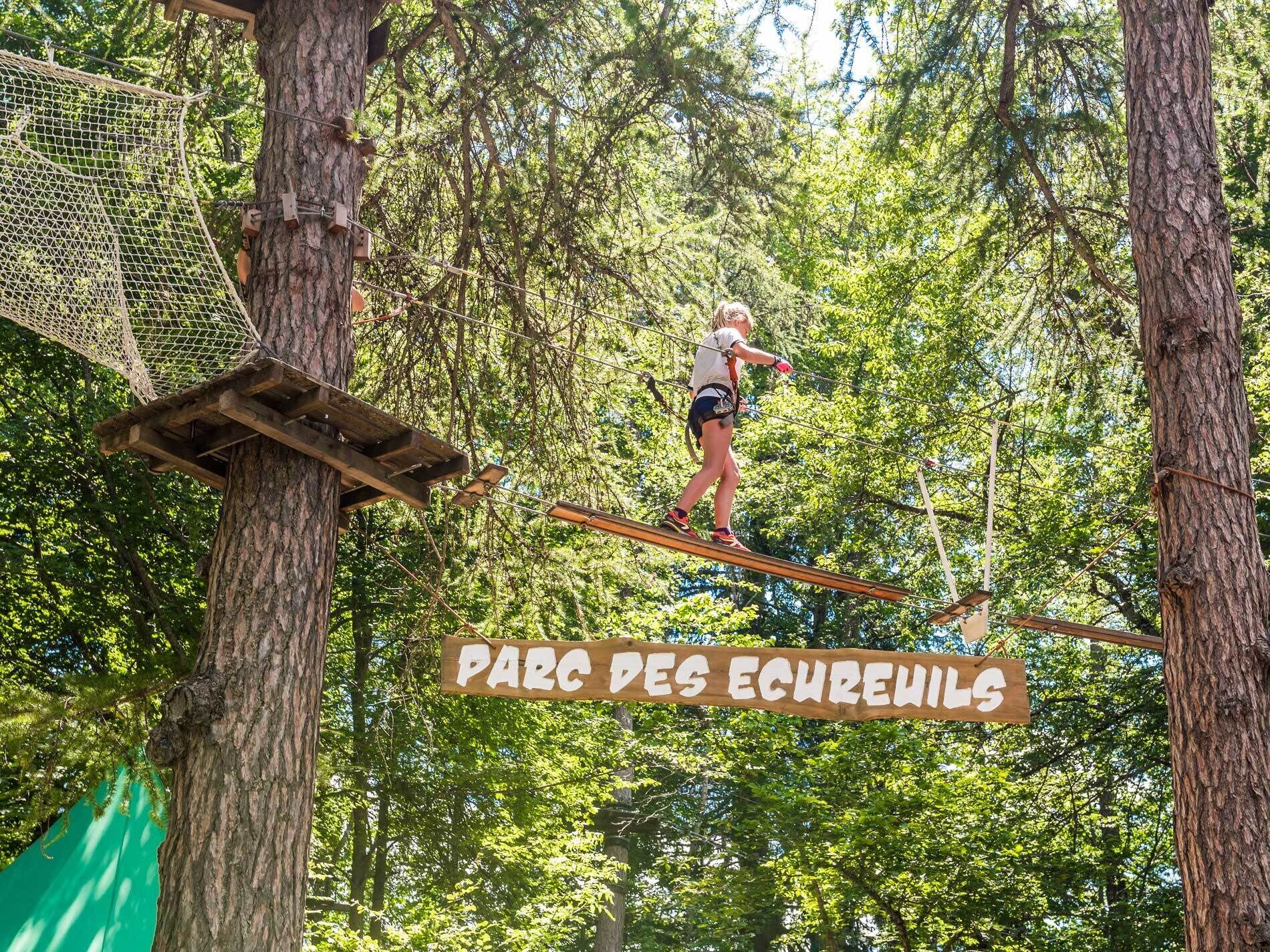 Une personne se trouve sur un pont de corde, dans une zone boisée, et un panneau indique : « PARC DES ÉCUREUILS ».