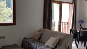A living room with a sofa, a small table, and a lamp. There is a window with a view of trees and a balcony. - Résidence Les Blanches Provençales (Selonnet)