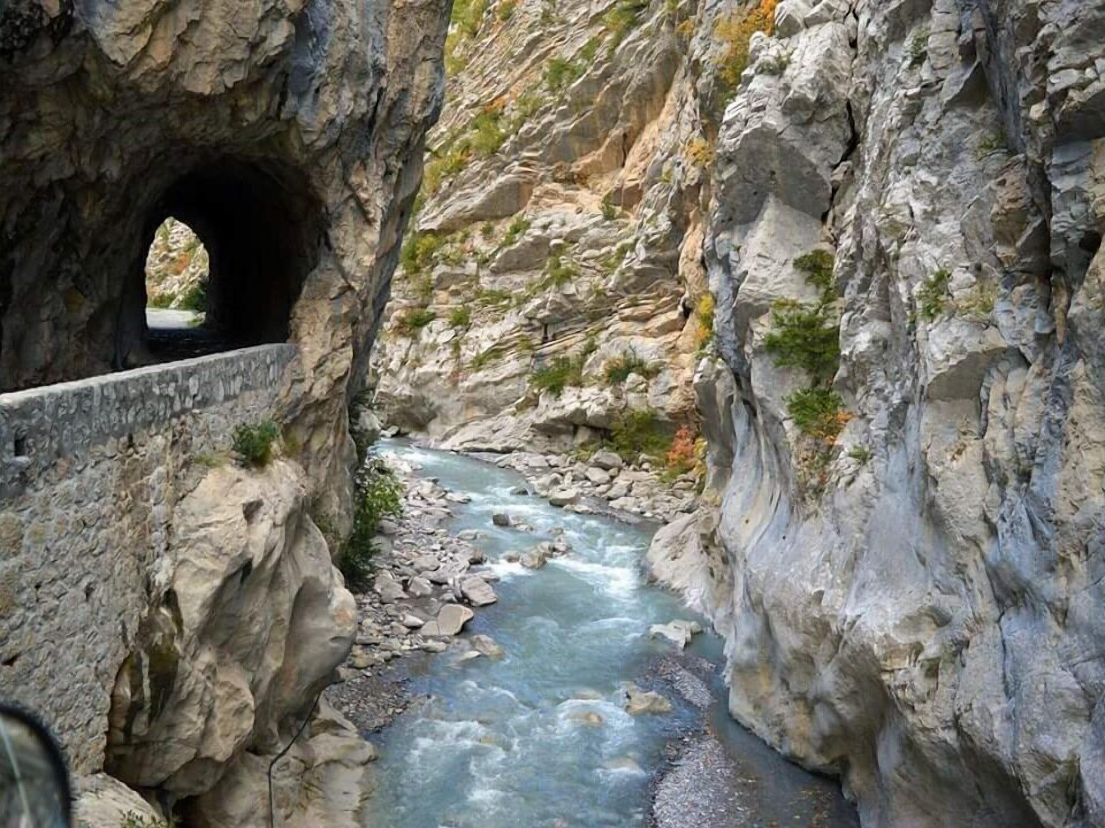 A narrow canyon with a flowing river, rocky walls, and a stone bridge.