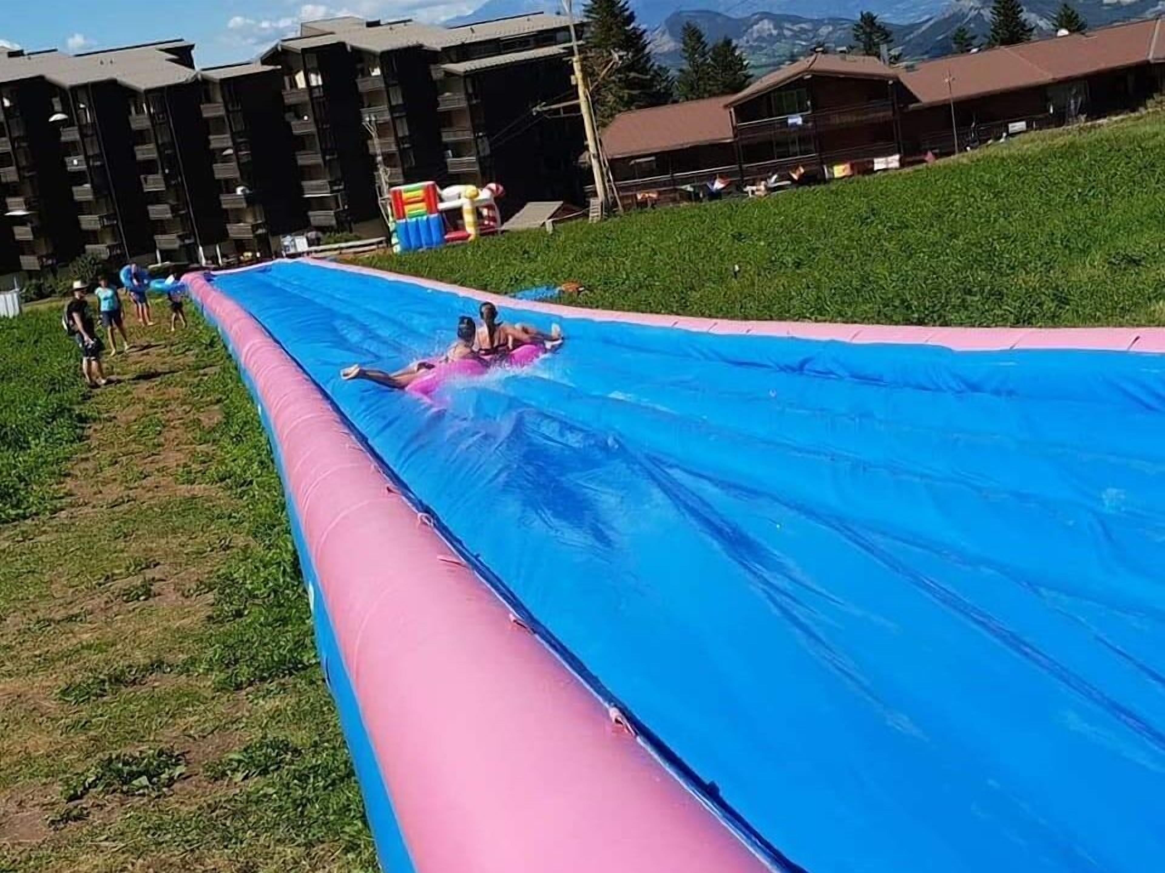 Un toboggan aquatique aux couleurs rose et bleu, entouré de zones verdoyantes et avec des bâtiments en arrière-plan.