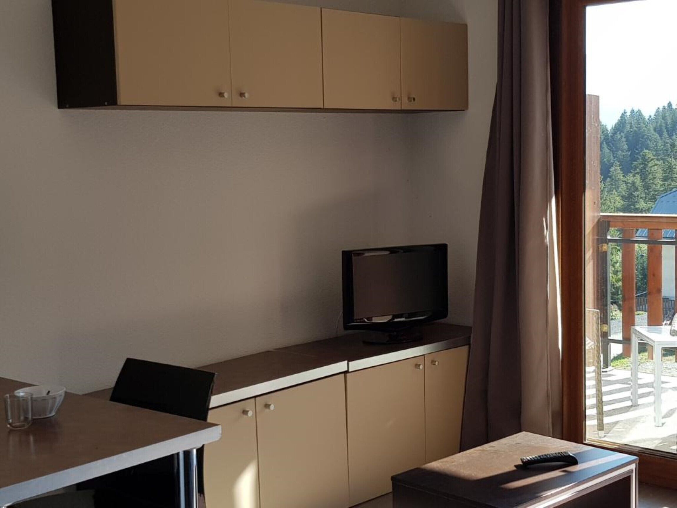 Une pièce avec un téléviseur posé sur un meuble, une table avec une chaise, et une vue sur une forêt par la fenêtre.