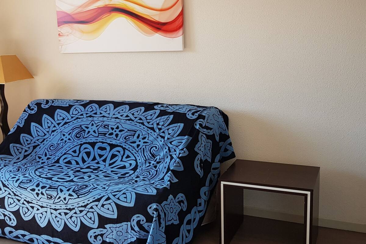 A room with a patterned blanket on a chair, a small table, and a wall with a colorful abstract painting.