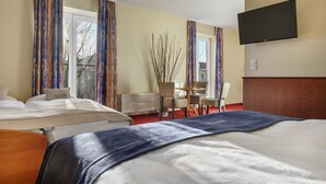 A hotel room with two beds, a TV mounted on the wall, a dining table with chairs, and a window with curtains.