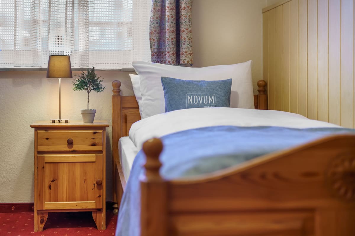 A neatly made bed with a blue pillow labeled "NOVUM," a wooden nightstand with a lamp, and a small potted plant.