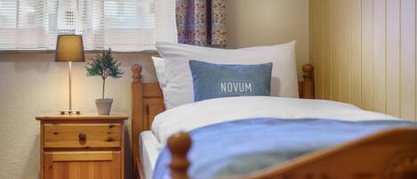 Cama bem arrumada com travesseiro azul com a etiqueta "NOVUM", mesa de cabeceira de madeira com luminária e uma pequena planta em vaso.
