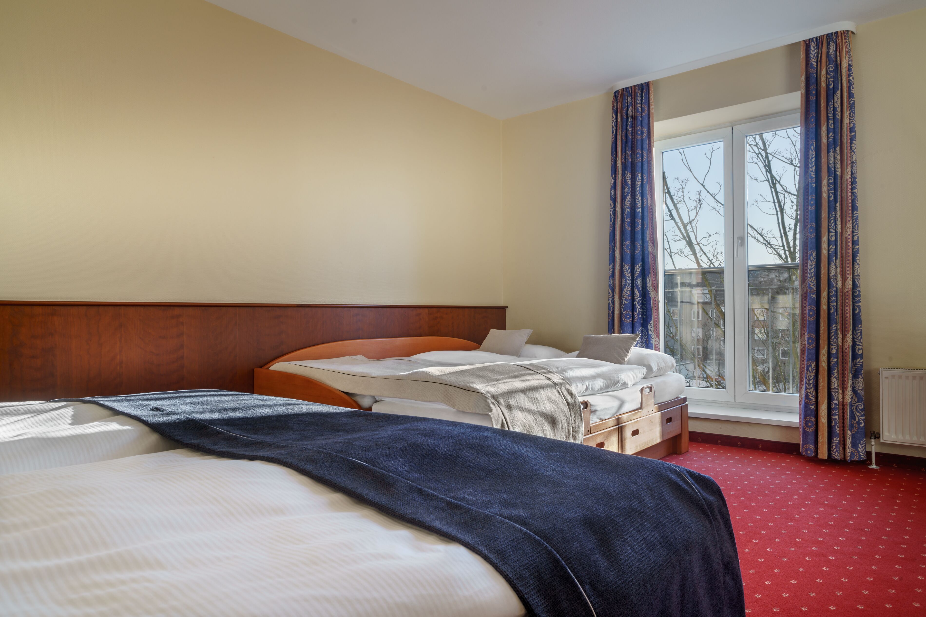A hotel room with a large bed, a smaller bed, a window with curtains, and a radiator.