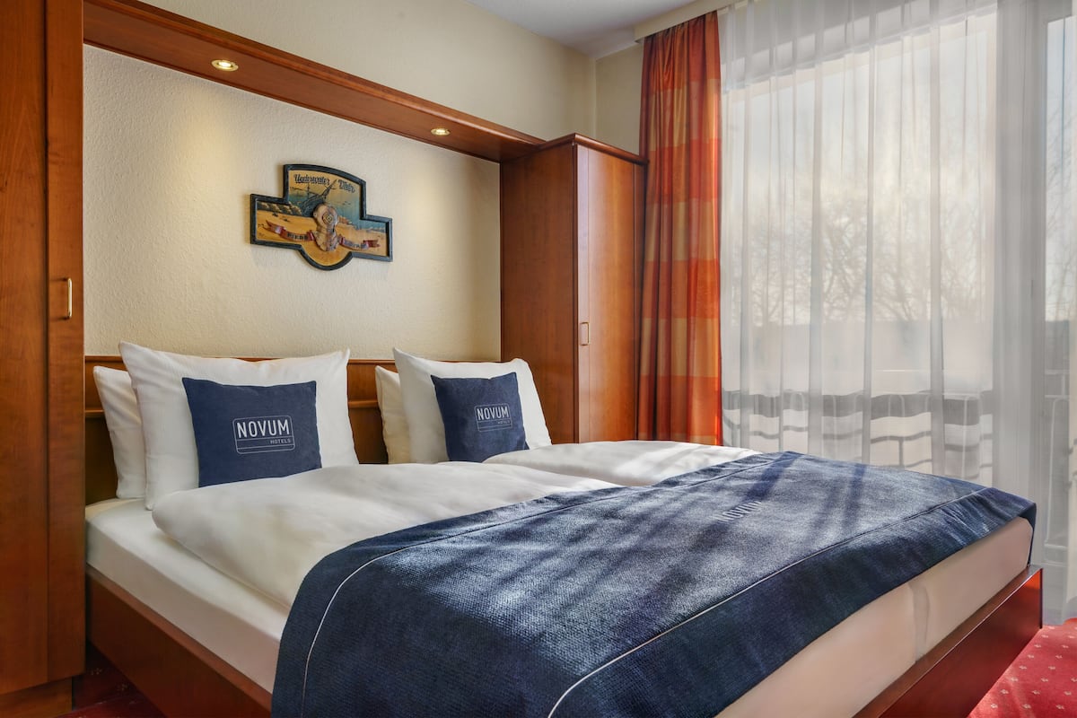 A hotel room with a bed, two pillows with 'Novum Hotels' branding, a wooden headboard, a wall clock, and a window with sheer curtains.