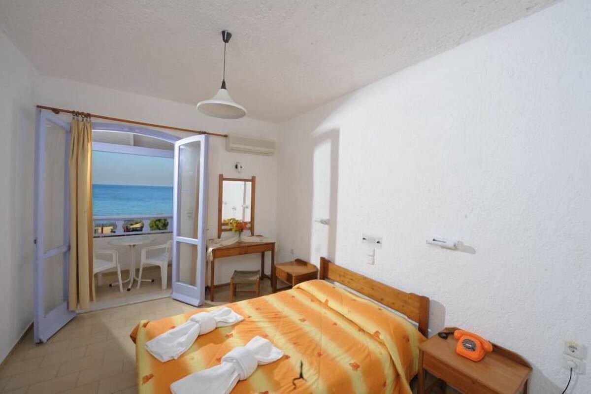 A bedroom with a bed, a desk, a chair, a telephone, and a view of the sea through an open door.