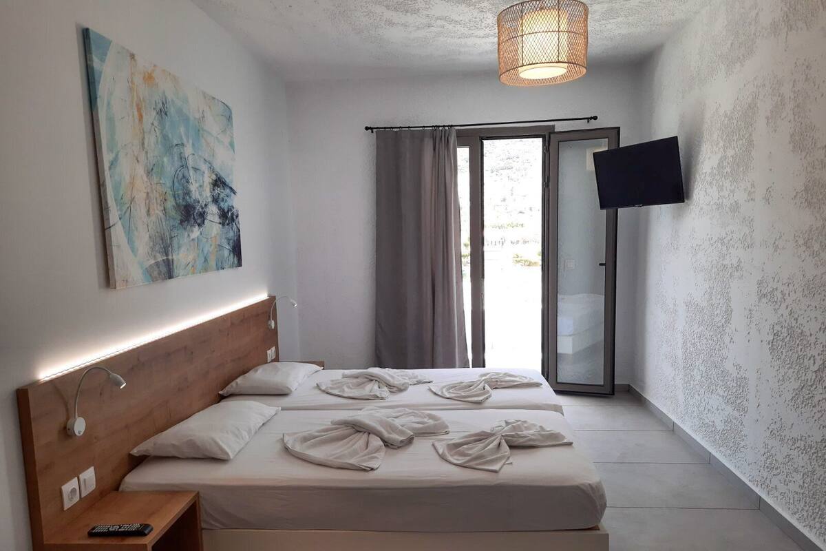 A modern bedroom with a wooden headboard, a bedside table with a remote control, a large bed with white linens, a wall-mounted TV, and a sliding glass door leading to an outdoor area.