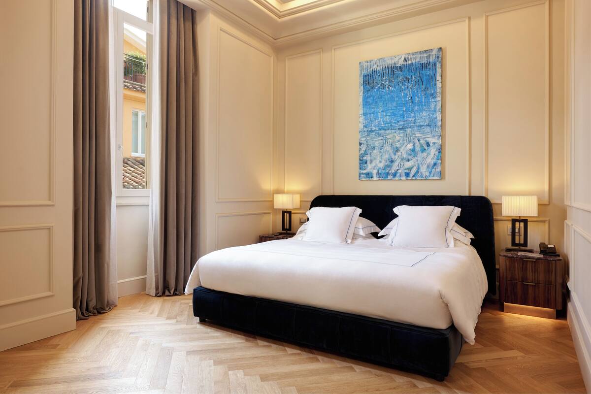A large bed with white linens, two bedside tables with lamps, a large abstract painting on the wall, and a view of a balcony through a window with curtains.