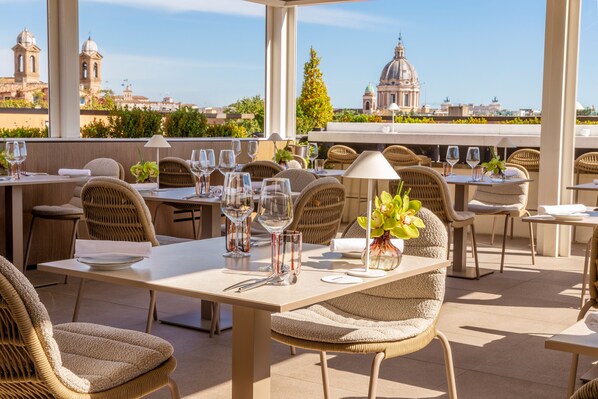 An outdoor dining area with wicker furniture, tables set with wine glasses, and a view of historic buildings.