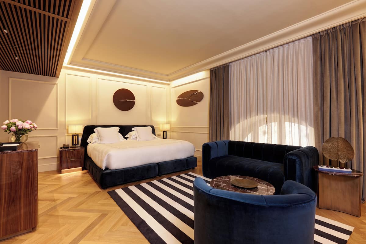 A modern hotel room with a large bed, a dark blue sofa, a wooden side table, and a striped rug.