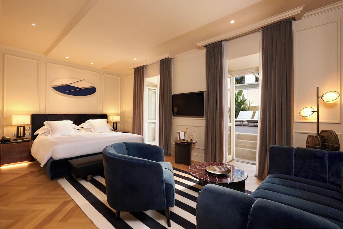 A modern hotel room with a large bed, two blue armchairs, a small coffee table, and a TV.