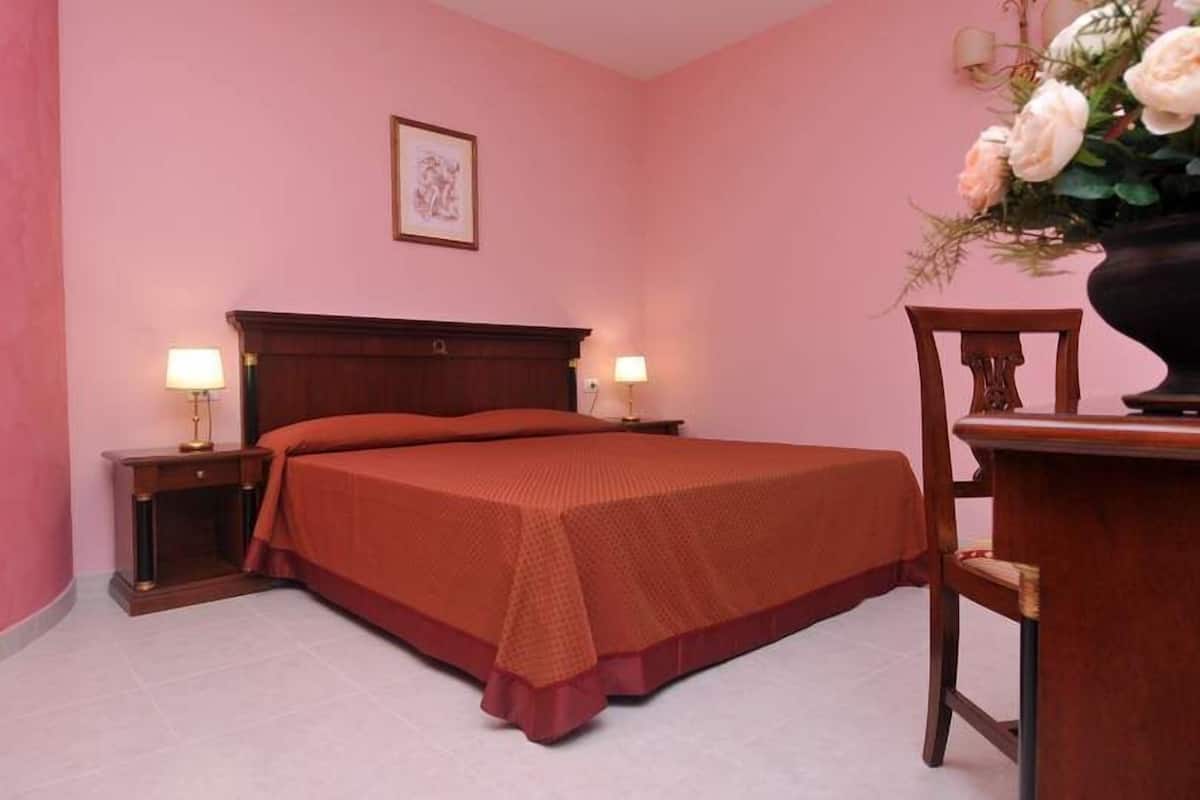 A bedroom with a large bed, two bedside tables with lamps, a wooden chair, and a vase with flowers.