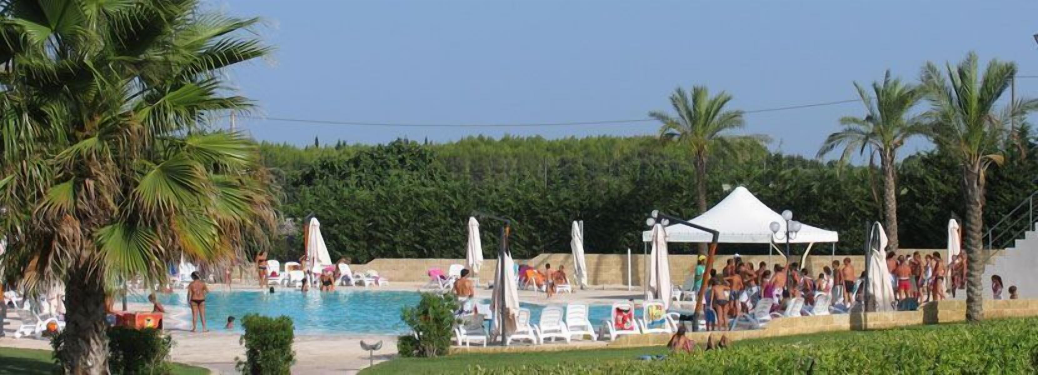 A pool area with lounge chairs and umbrellas, surrounded by palm trees and a white tent.