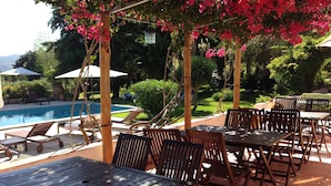 Un patio exterior con mesas y sillas de madera, una piscina y exuberante follaje.