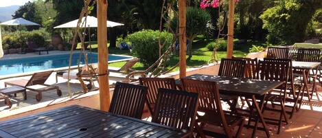 An outdoor patio with wooden tables and chairs, a pool, and lush greenery.