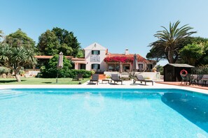 Piscina com espreguiçadeiras e guarda-sóis, rodeada por um jardim e um edifício com telhado de telhas vermelhas.