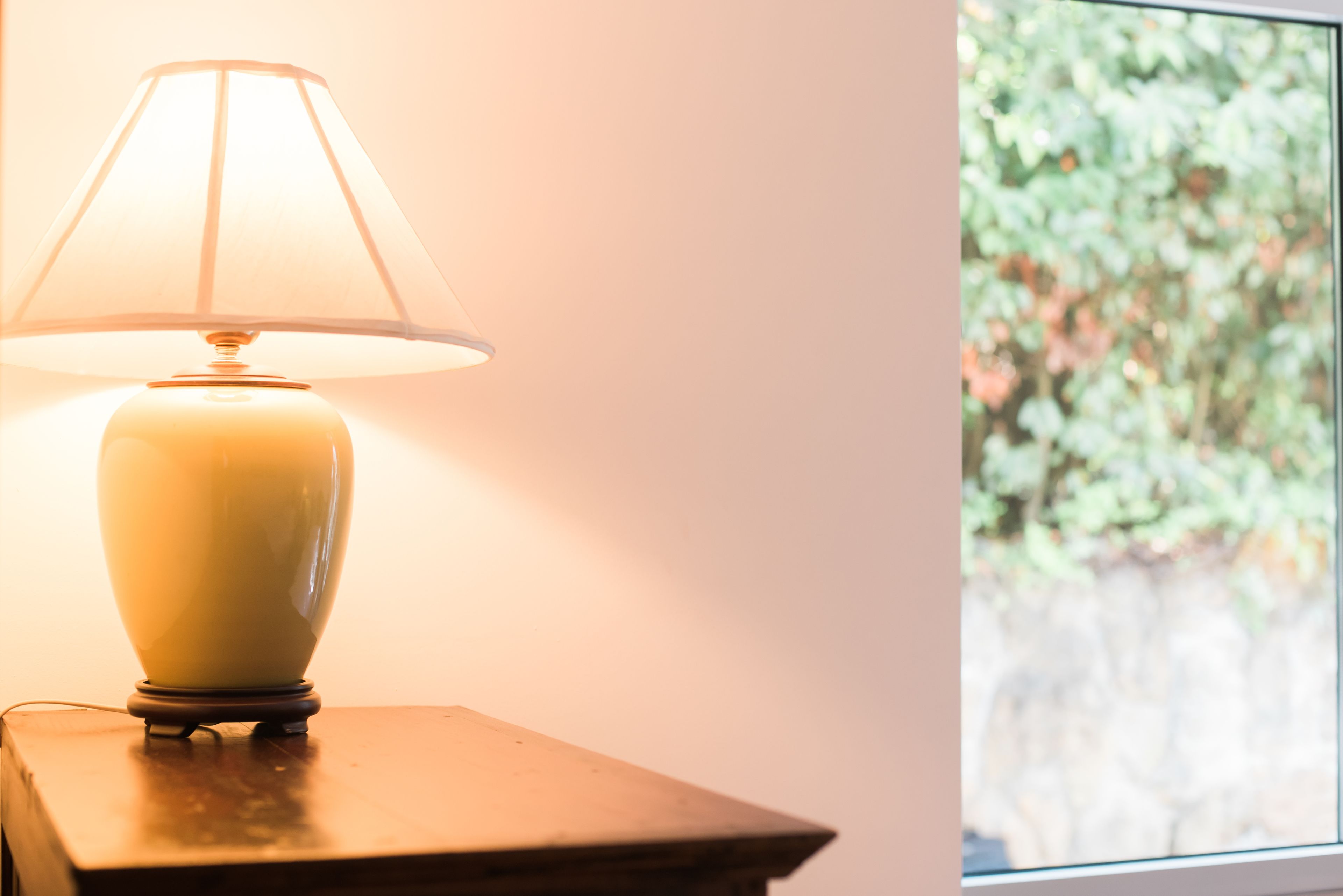 A table lamp with a white shade on a wooden table next to a window with a view of greenery.