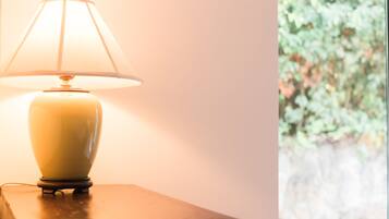 A table lamp with a white shade on a wooden table next to a window with a view of greenery.