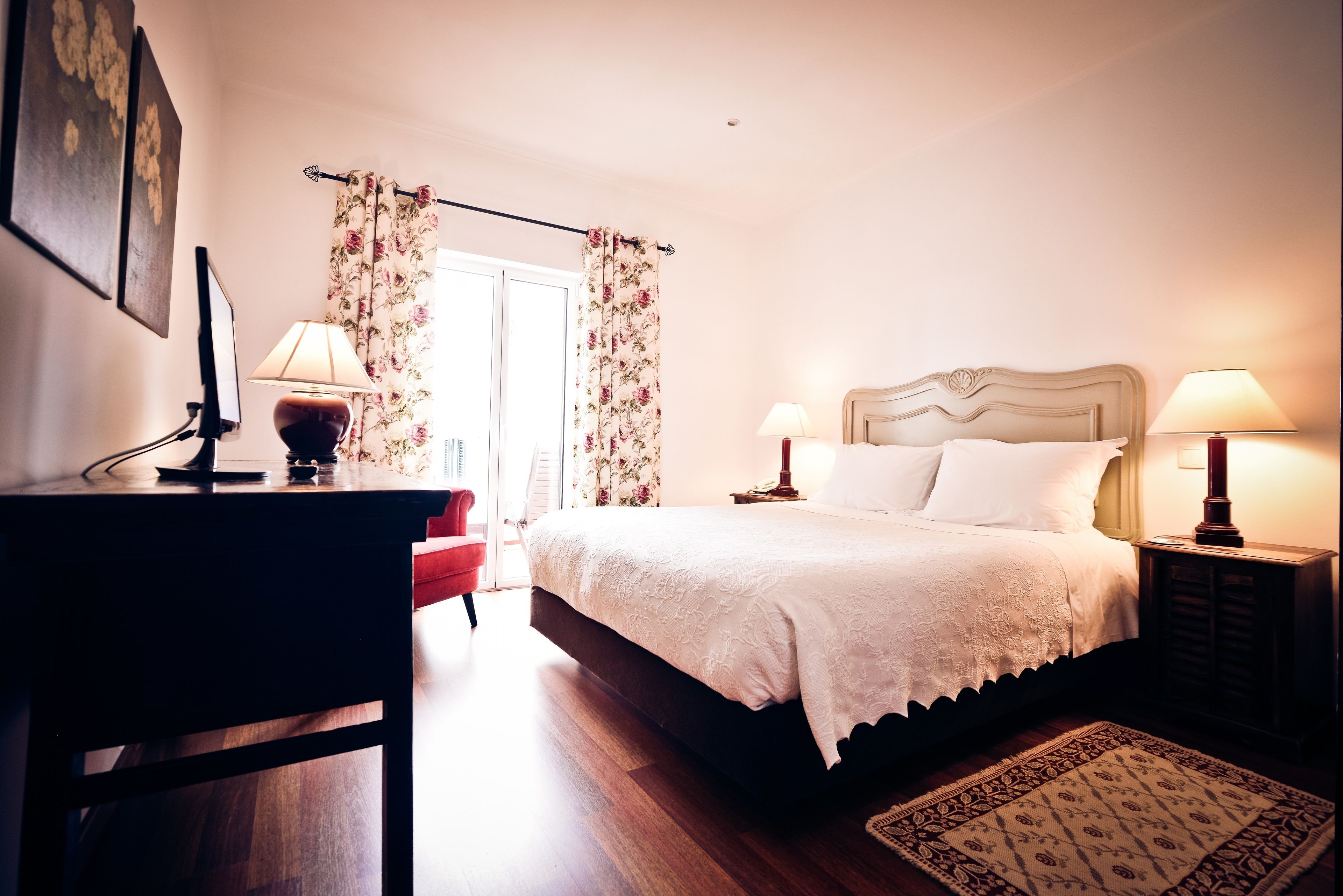 A bedroom with a bed, two bedside lamps, a desk with a lamp, a chair, a rug, and a door with curtains.