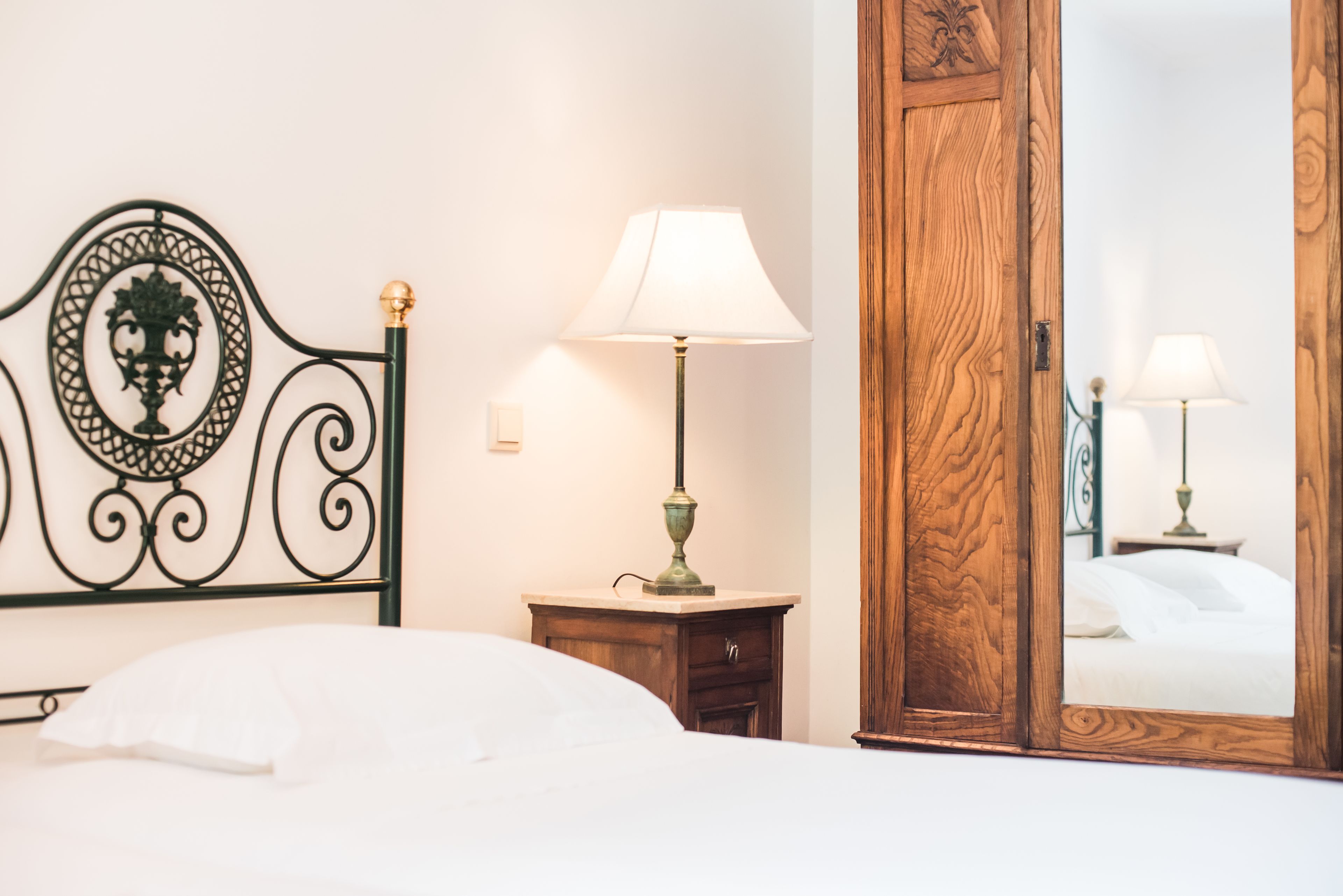Una cama bien hecha con cabecera, una lámpara de noche, un armario de madera y un espejo que refleja la habitación.