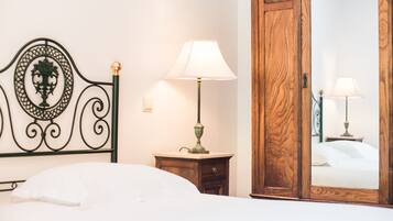 Una cama bien hecha con cabecera, una lámpara de noche, un armario de madera y un espejo que refleja la habitación.