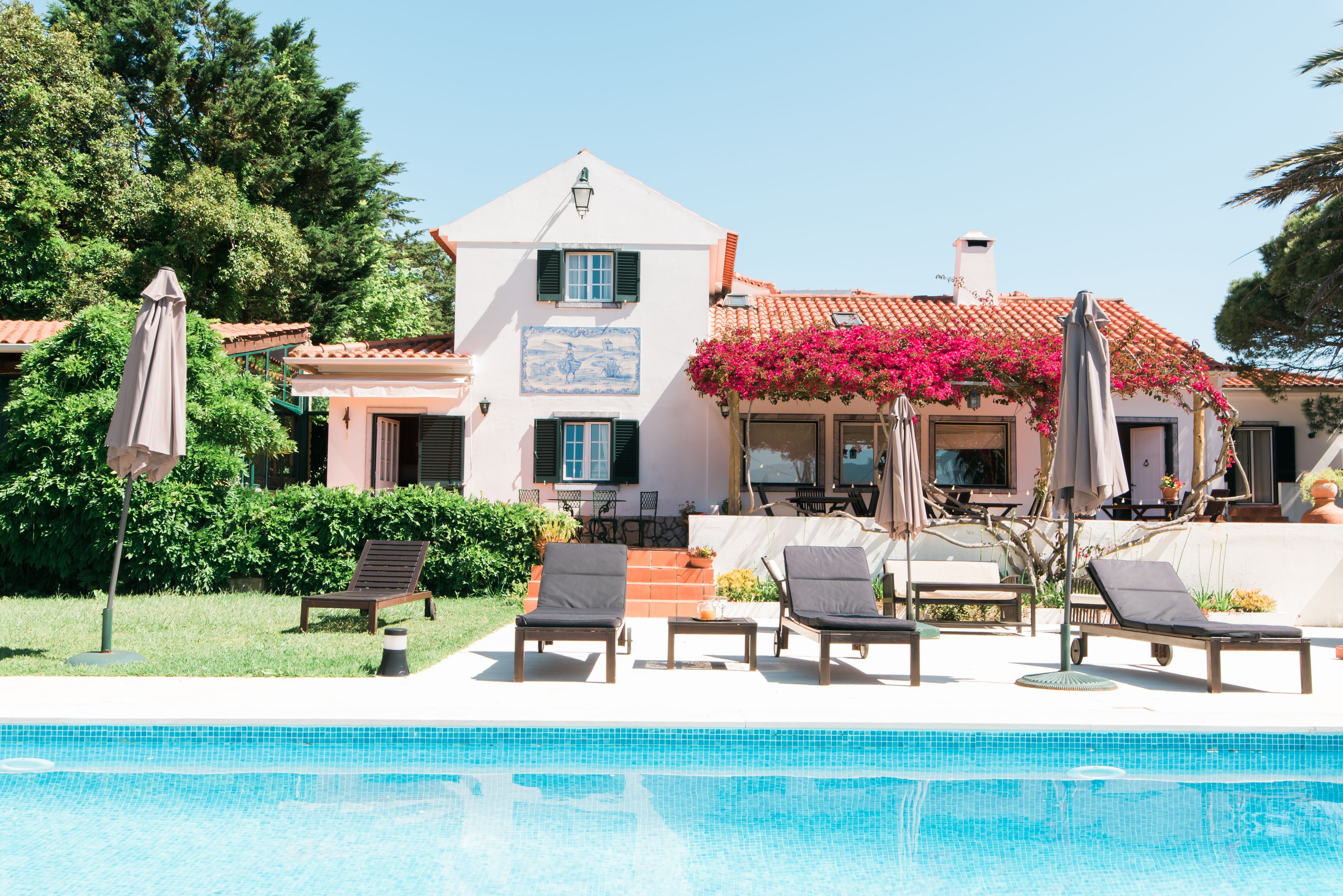 A pool area with lounge chairs and umbrellas, a house with a tiled roof, and a garden with bushes and flowers.