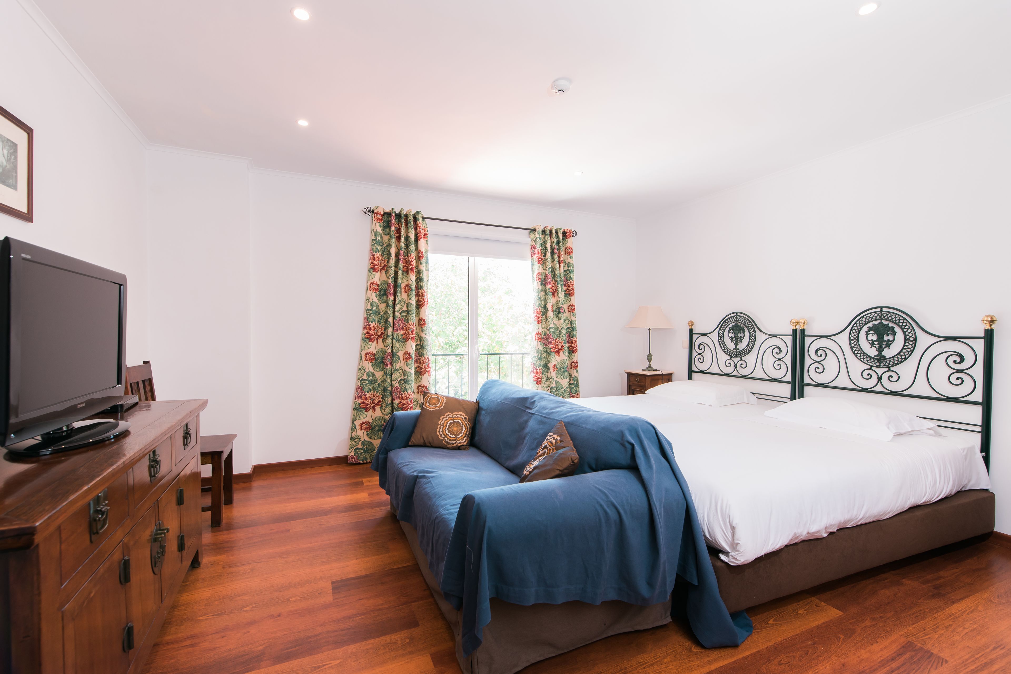 A bedroom with a bed, a television on a wooden dresser, and a window with floral curtains.