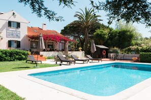 Piscina com espreguiçadeiras e guarda-sóis, rodeada por vegetação e um edifício com telhado em azulejos.