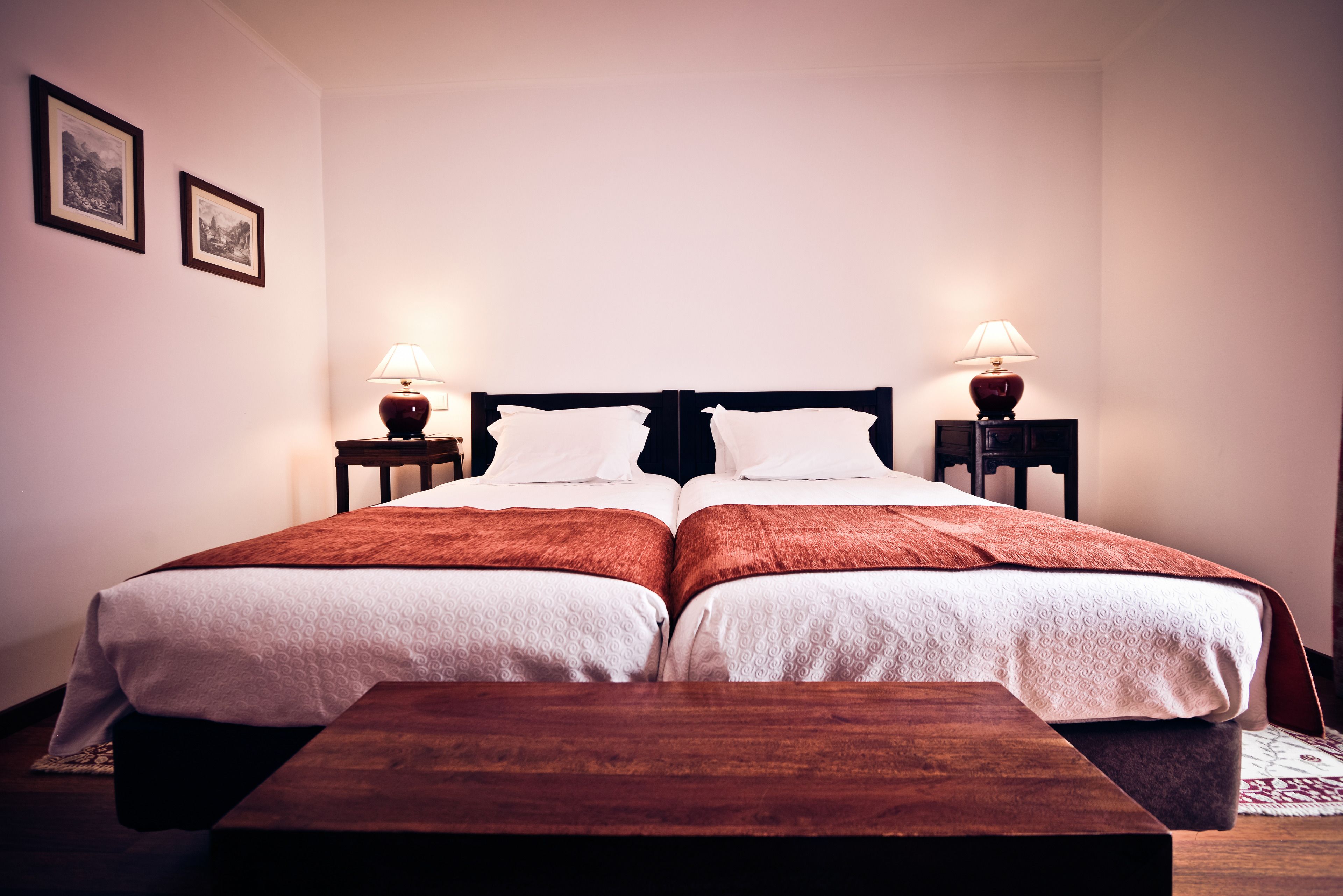 A double bed with two nightstands, each with a lamp, in a room with plain walls and a wooden bench.