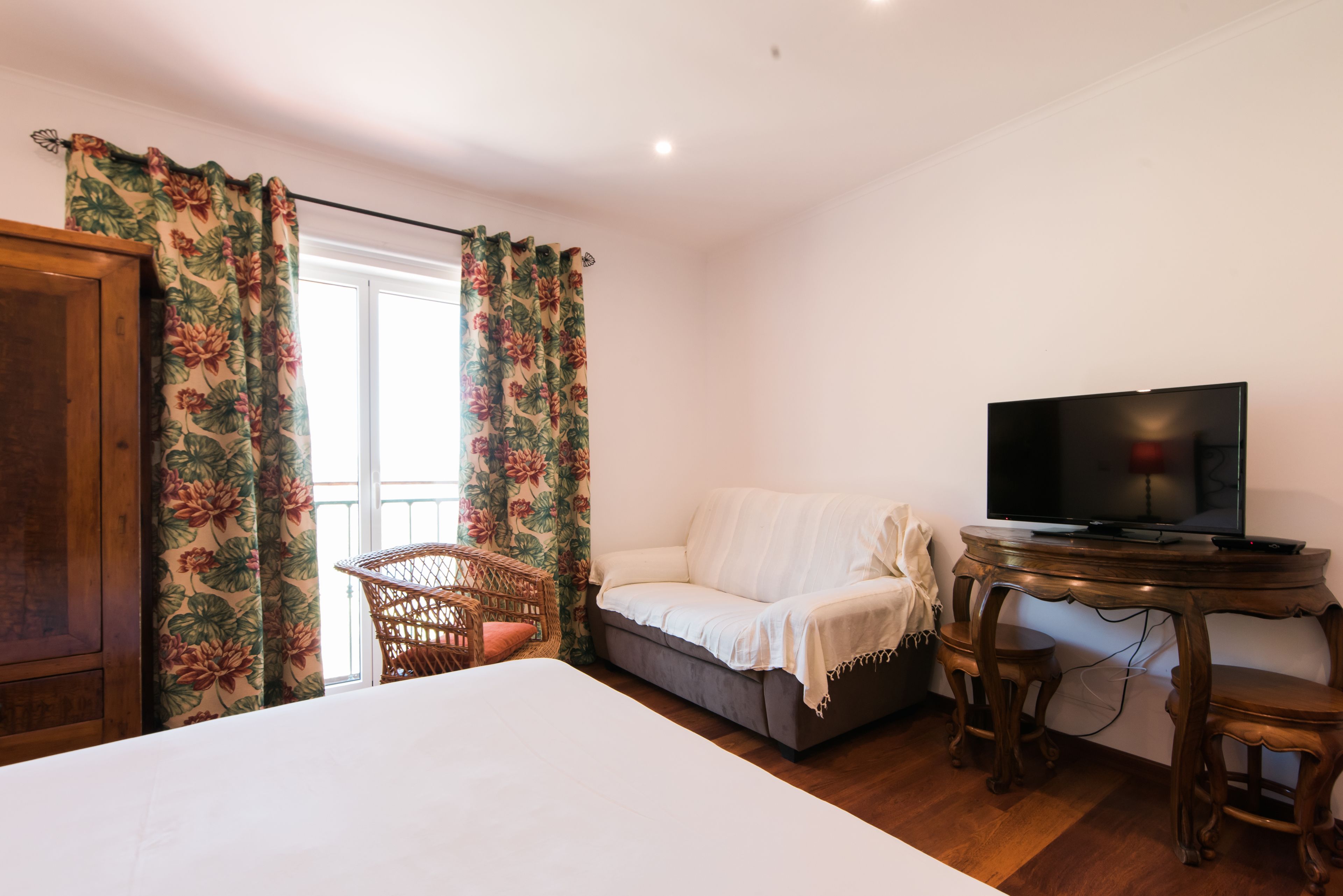 Une chambre à coucher avec un lit, un canapé, un téléviseur posé sur une table en bois et un fauteuil en osier.