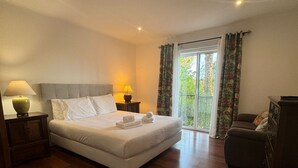 Un dormitorio con una cama grande, suelo de madera, una mesita de noche con lámpara, una silla y vistas a zonas verdes a través de la ventana.