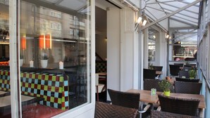 An outdoor cafe area with wicker chairs and tables, a checkered bench, and a glass wall with a view of the street. - Hotel La Terrasse (De Panne)