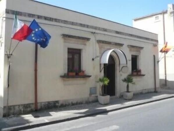 A building with a white facade, a small arched entrance, and two potted plants on the ground floor. - Albergo La Fontanella (Noto)