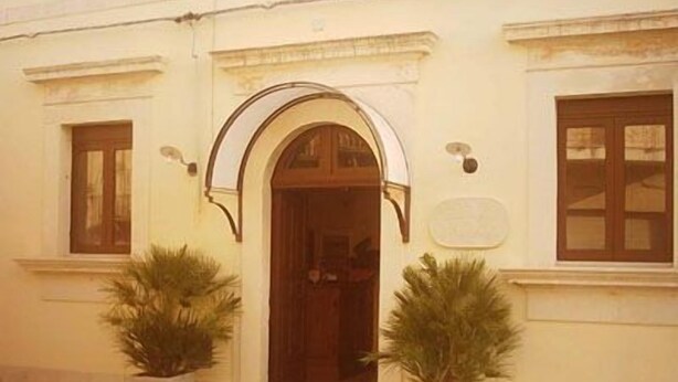 A building with a central arched entrance, two potted plants on either side, and a small green area in front. - Albergo La Fontanella (Noto)