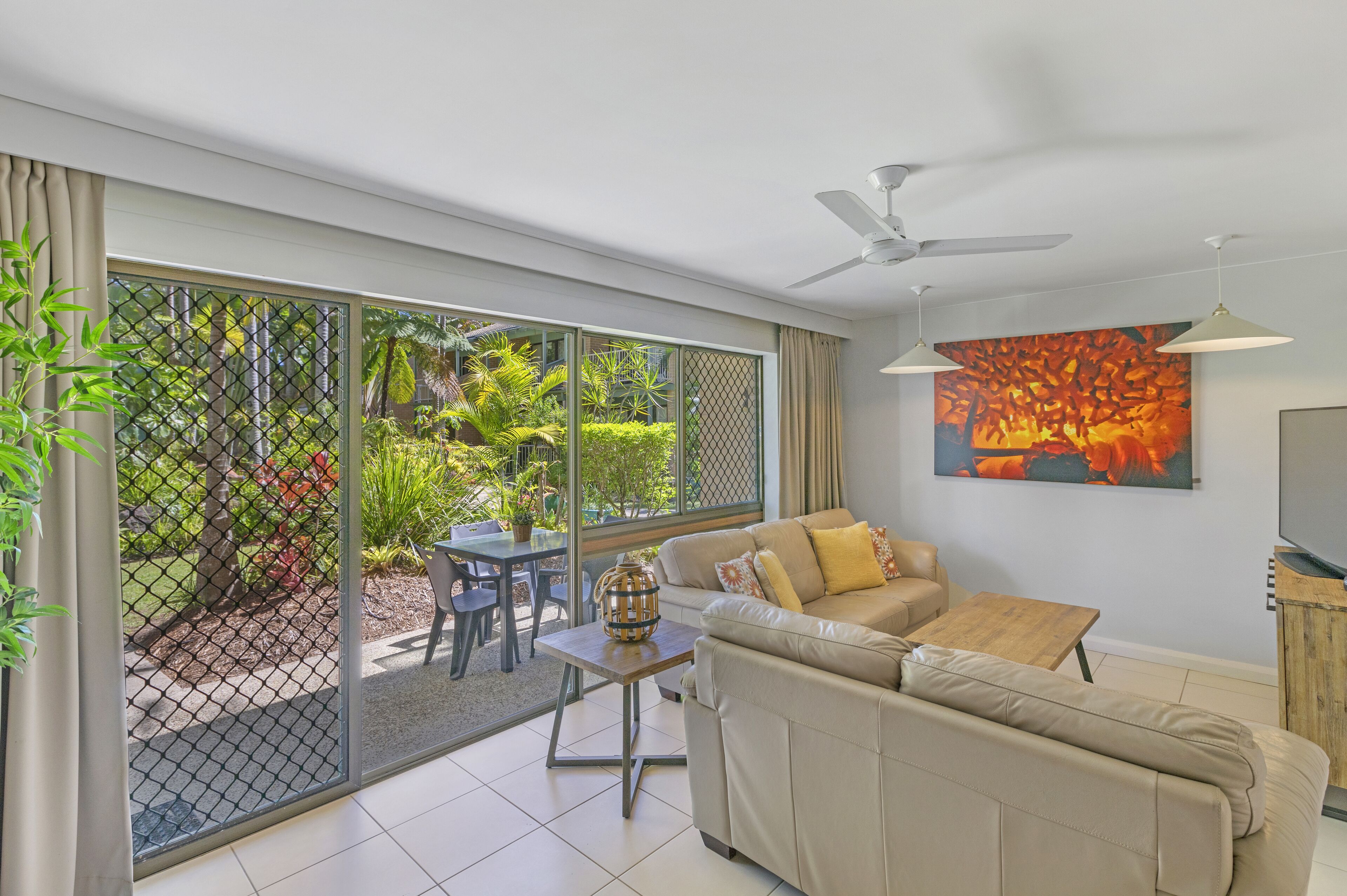 A modern living room with a sofa, a coffee table, and a TV. There is a sliding glass door leading to an outdoor area with plants and a table.