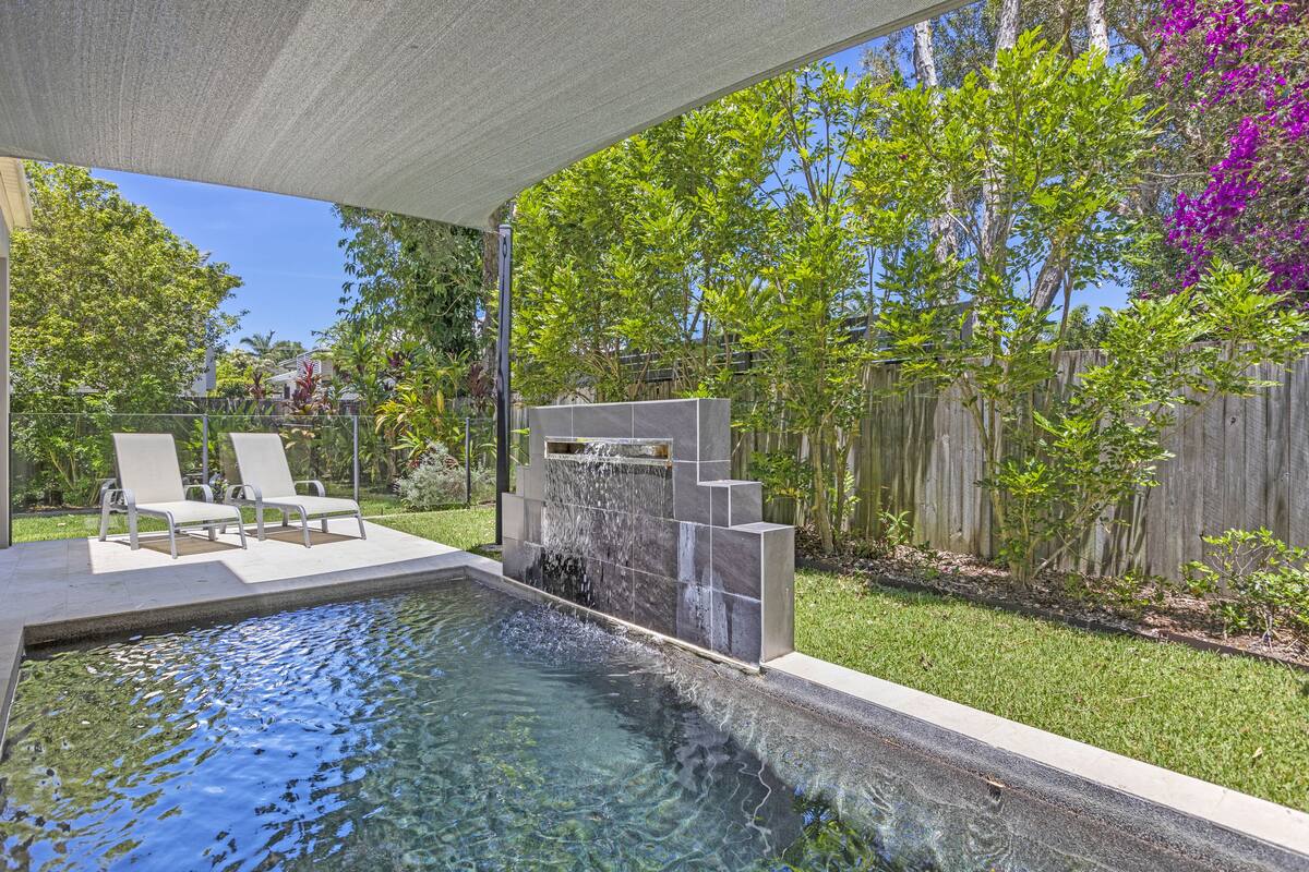 A swimming pool with a waterfall feature, surrounded by a patio with chairs and a wooden fence.