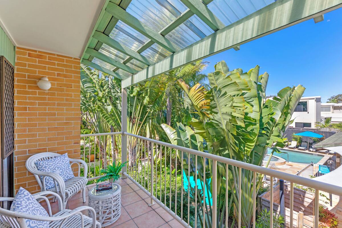 A balcony with wicker furniture, a potted plant, and a view of a swimming pool and tropical plants.