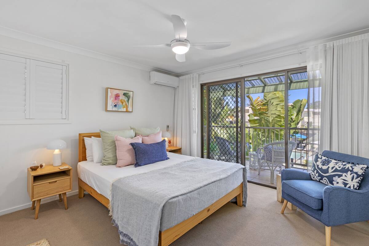 A bedroom with a bed, a nightstand, a chair, a ceiling fan, and a view of the outdoors.