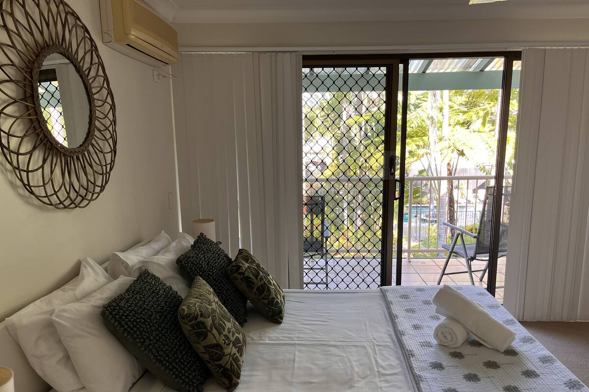 A hotel room with a bed, white curtains, a balcony with a metal railing, and a view of greenery outside.
