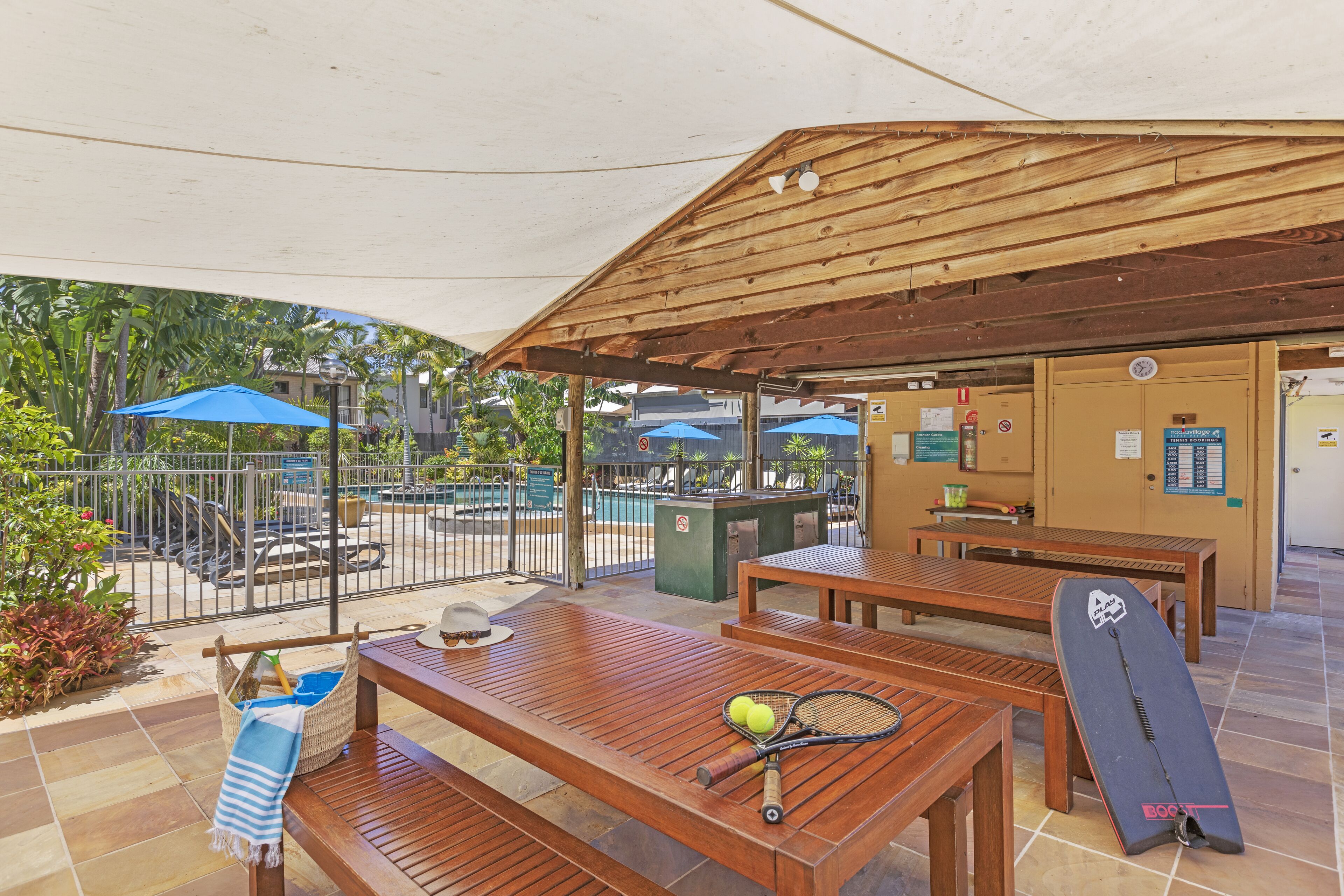 An outdoor seating area with wooden tables and benches, a pool in the background, and a covered section with a wooden roof.