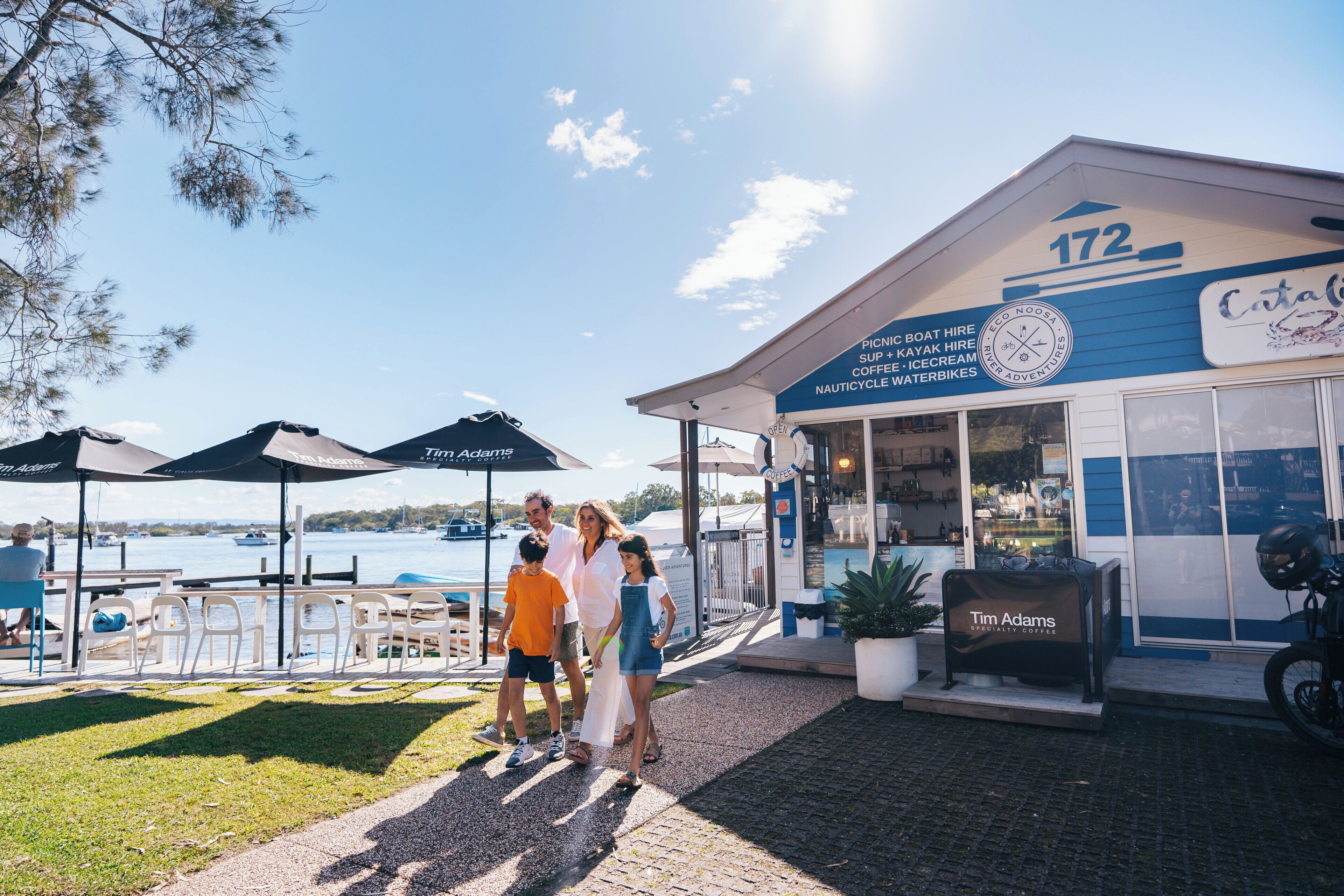 A waterfront restaurant with outdoor seating and umbrellas, a sign for Tim Adams coffee, and a view of the water.