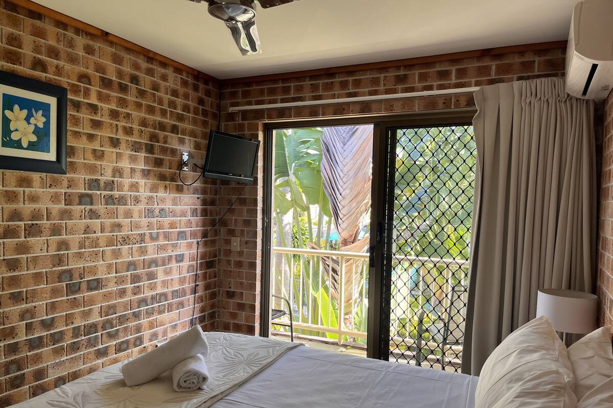 A hotel room with a brick wall, a bed with white bedding, a TV mounted on the wall, and a sliding glass door leading to an outdoor area with greenery.