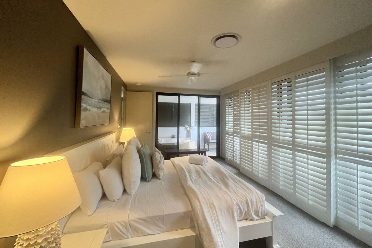 A bedroom with a bed, a bedside lamp, a window with blinds, and a view of a balcony.