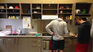 A kitchen with two people standing at the counter, a stove, and various kitchen items on shelves. - Sydney City Hostel (Haymarket)