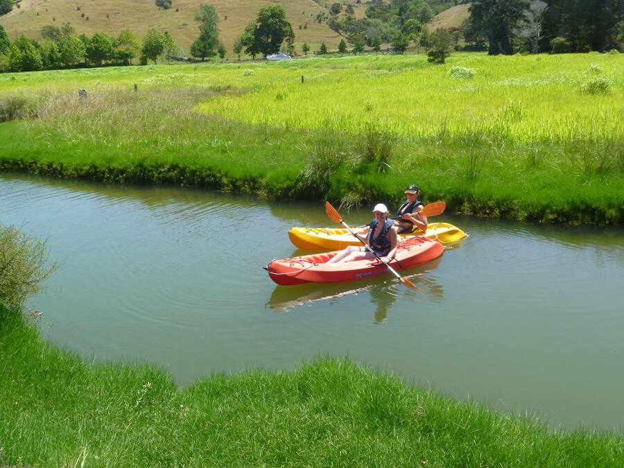 Deux personnes pratiquent le kayak sur une rivière paisible, entourée de champs verdoyants et d’arbres.
