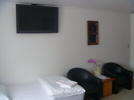 Una habitación pequeña y sencilla con una cama, dos sillas negras, una mesita, un cuadro en la pared y un televisor colgado sobre la cama.