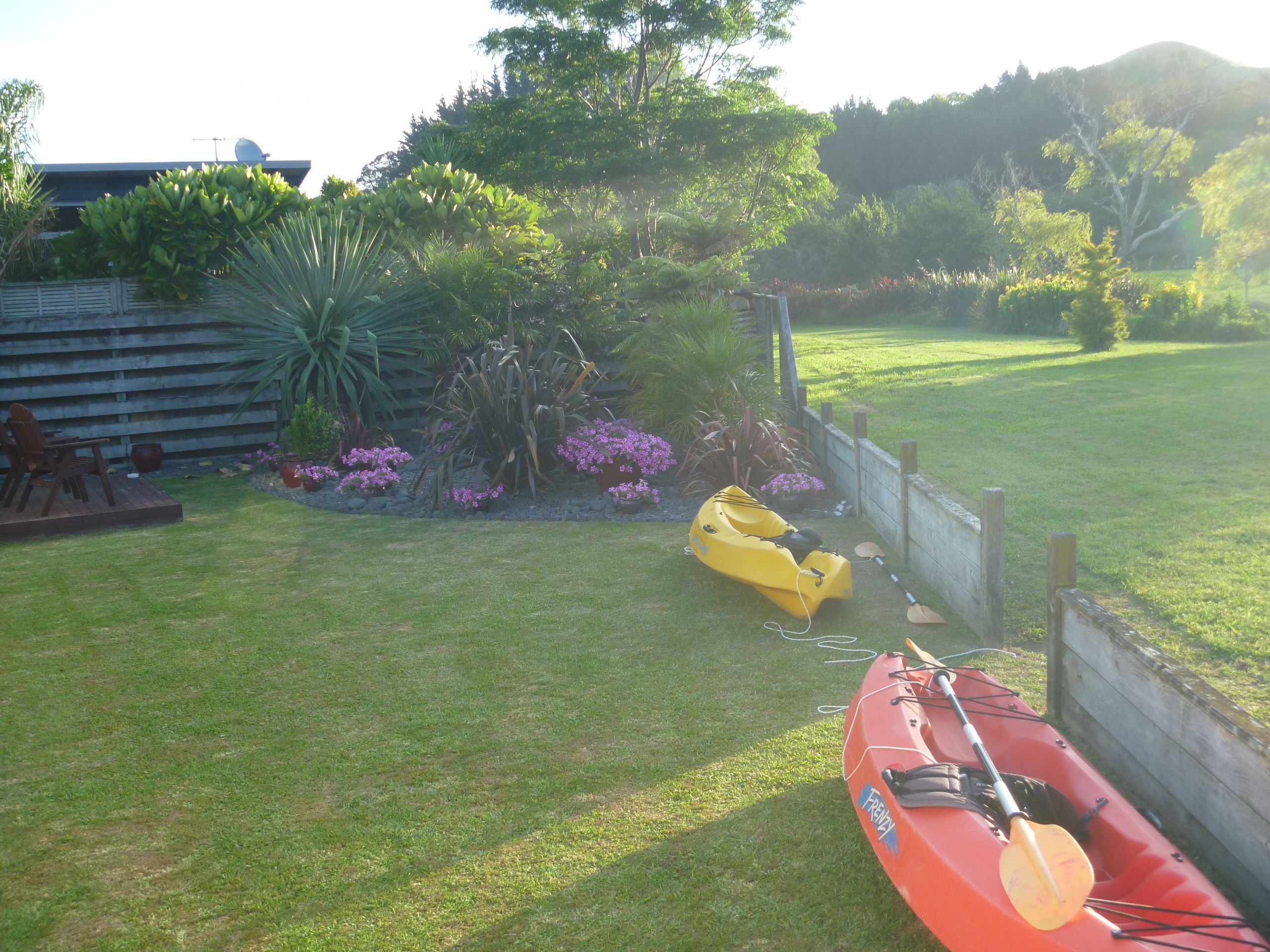 A grassy yard with a yellow kayak and paddle, a wooden deck with chairs, and a variety of plants and flowers.