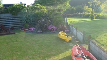 A grassy yard with a yellow kayak and paddle, a wooden deck with chairs, and a variety of plants and flowers.